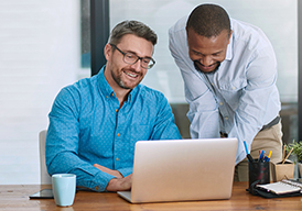  Man sitting at a conference table with another man standing next to him; both looking at a laptop computer