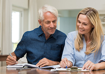 Older man with an open book and younger woman with a clipboard sitting at a table, looking at papers