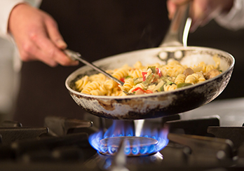 Hands holding a fry pan cooking food over a gas stove burner
