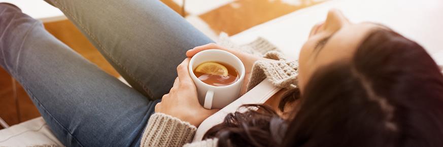 Woman at home wearing a sweater lounging comfortably on a couch drinking hot tea with lemon.